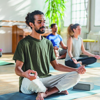 Foto eines Yoga Raums mit mehreren Personen, die im Schneidersitz sitzen. Im Vordergrund ist ein Mann zu sehen, er sitzt ebenfalls im Schneidersitz und sieht entspannt aus.