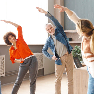 Gruppe von Erwachsenen führt gemeinsam eine seitliche Dehnübung in einem hellen Raum durch. Alle lächeln und bewegen sich synchron, um ihre Beweglichkeit zu fördern.